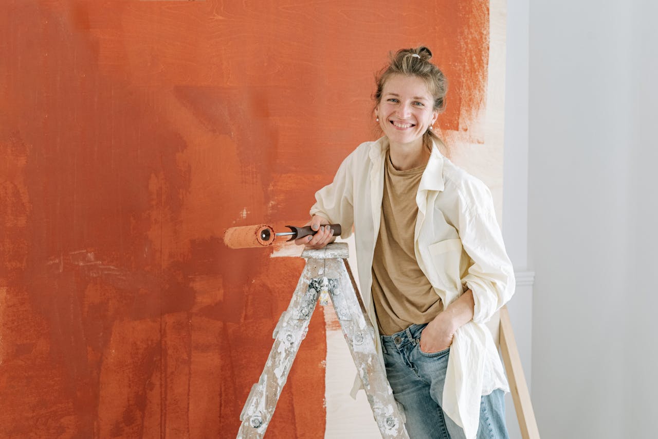 services-02 A cheerful woman in casual attire paints an orange wall while standing on a ladder indoors.